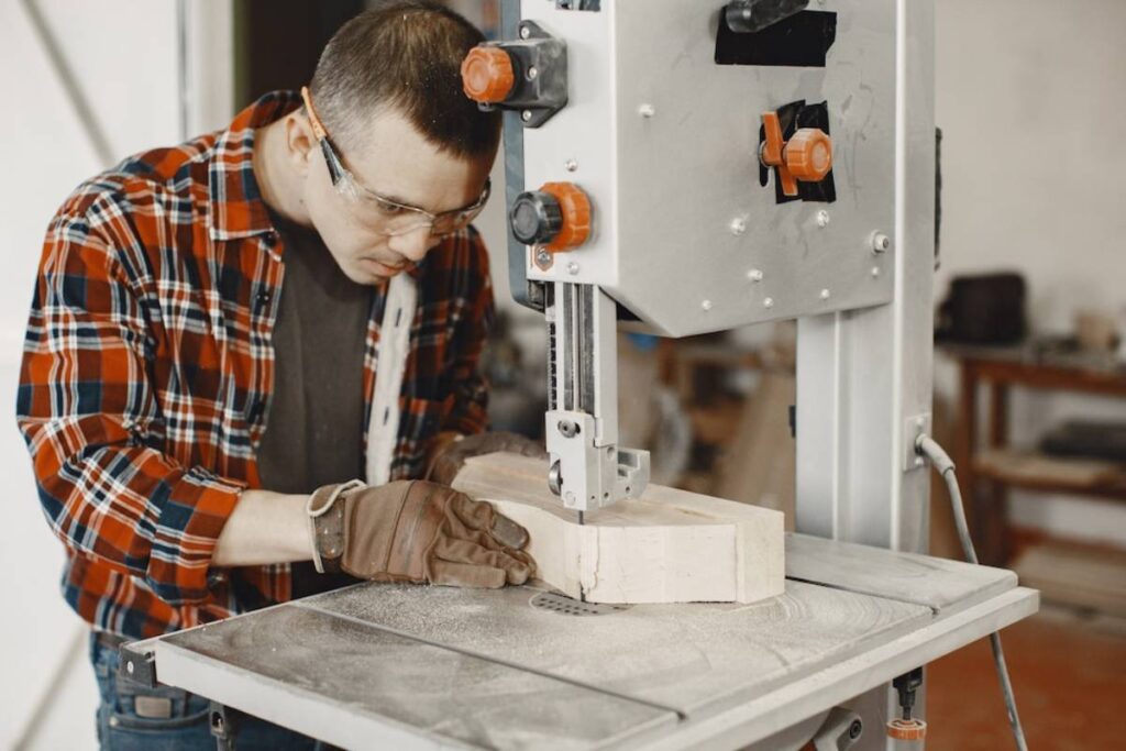 A person in a plaid shirt and gloves uses a bandsaw to cut a piece of wood in a workshop, surrounded by tools and sawdust.
