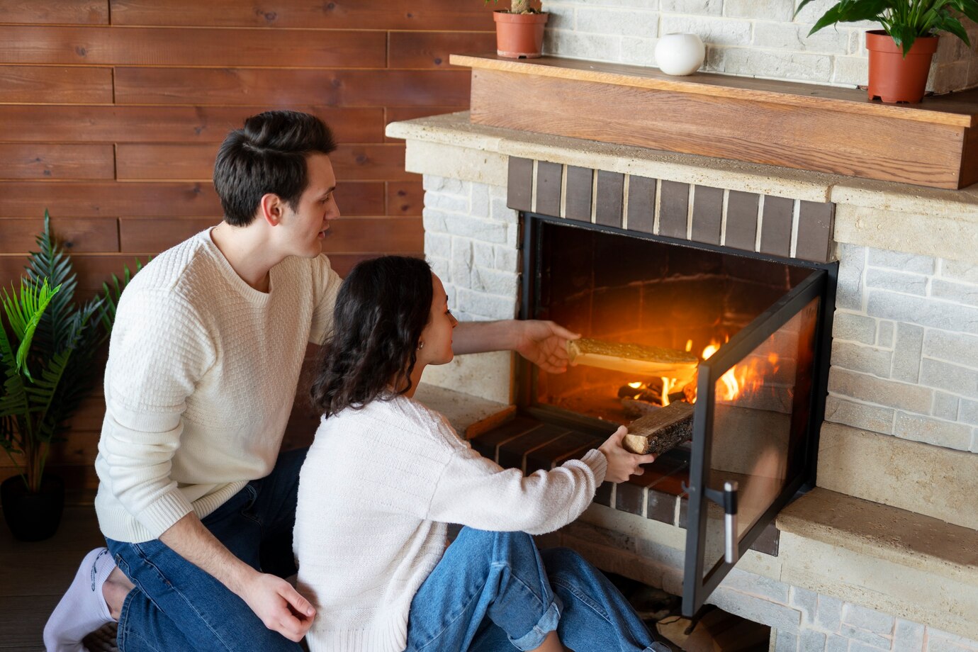 couple sits close to a cozy fireplace, adding logs to the flames