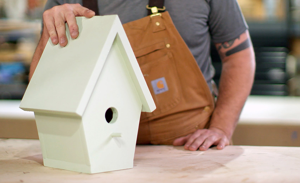 person in a gray shirt and brown overalls holds a mint green birdhouse
