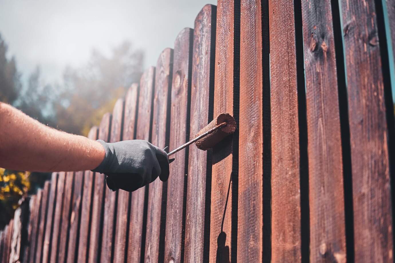 hand in a black glove paints a wooden fence with a paint roller