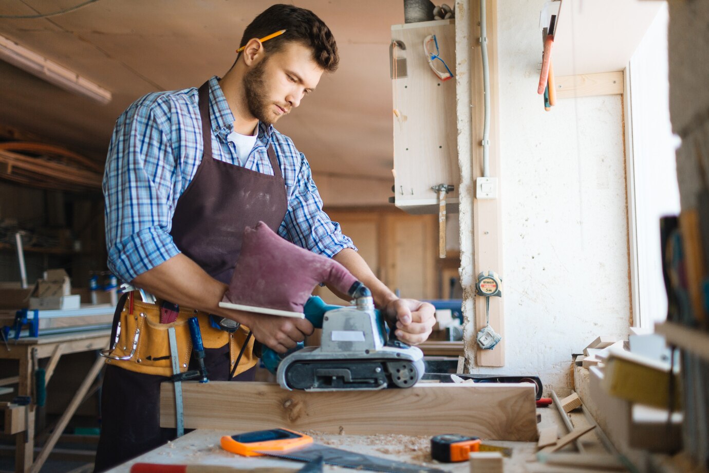 person in a checkered shirt and apron operates a belt sander on a wooden plank