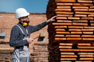 worker in a hard hat and ear protection inspects stacked wooden planks