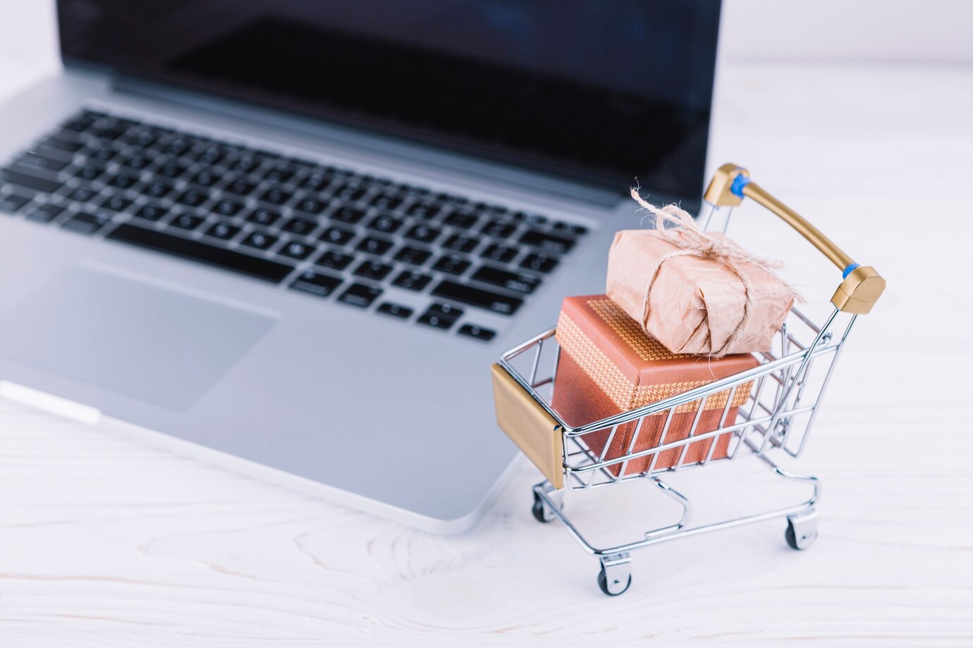 small shopping cart with wrapped gifts sits beside a laptop
