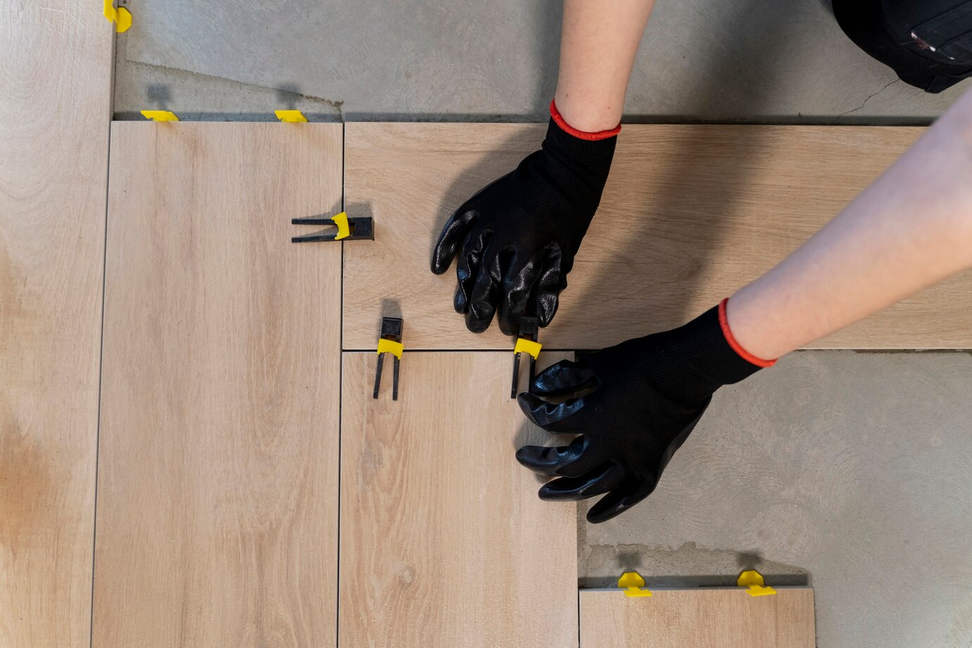 pair of hands wearing black gloves lays wooden tiles on a floor