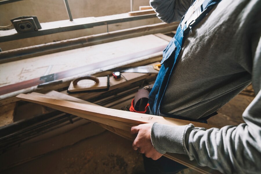 carpenter in a gray sweatshirt holds a wooden plank, surrounded by tools 