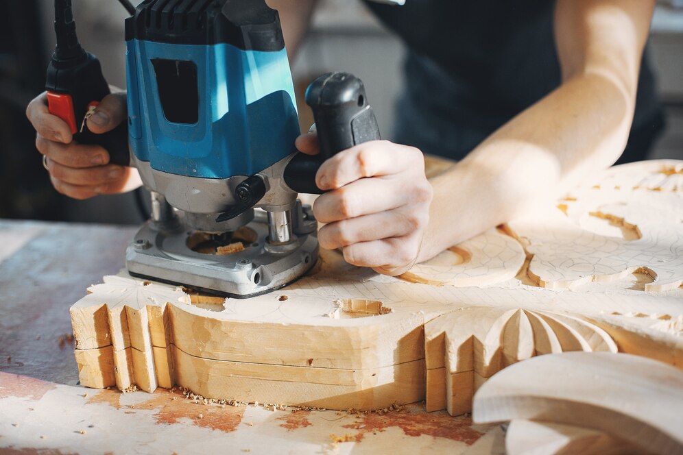person using a blue electric router on a wooden piece, shaping intricate carvings