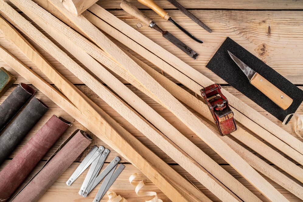 A flat lay of woodworking tools, including wooden strips, sandpaper, a plane, and various hand tools, on a wooden surface.