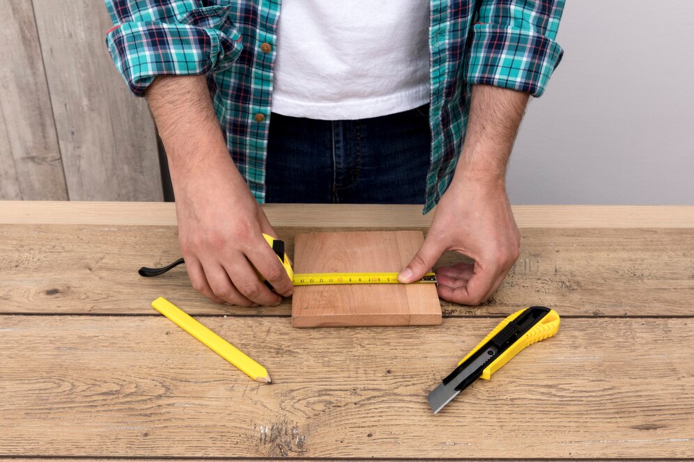 A person measuring a wooden block on a table, with measuring tape, yellow pencil, and utility knife beside them.