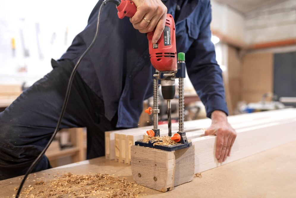 A carpenter uses a power drill on a wooden jig, with wood shavings scattered on the workbench in a workshop setting.