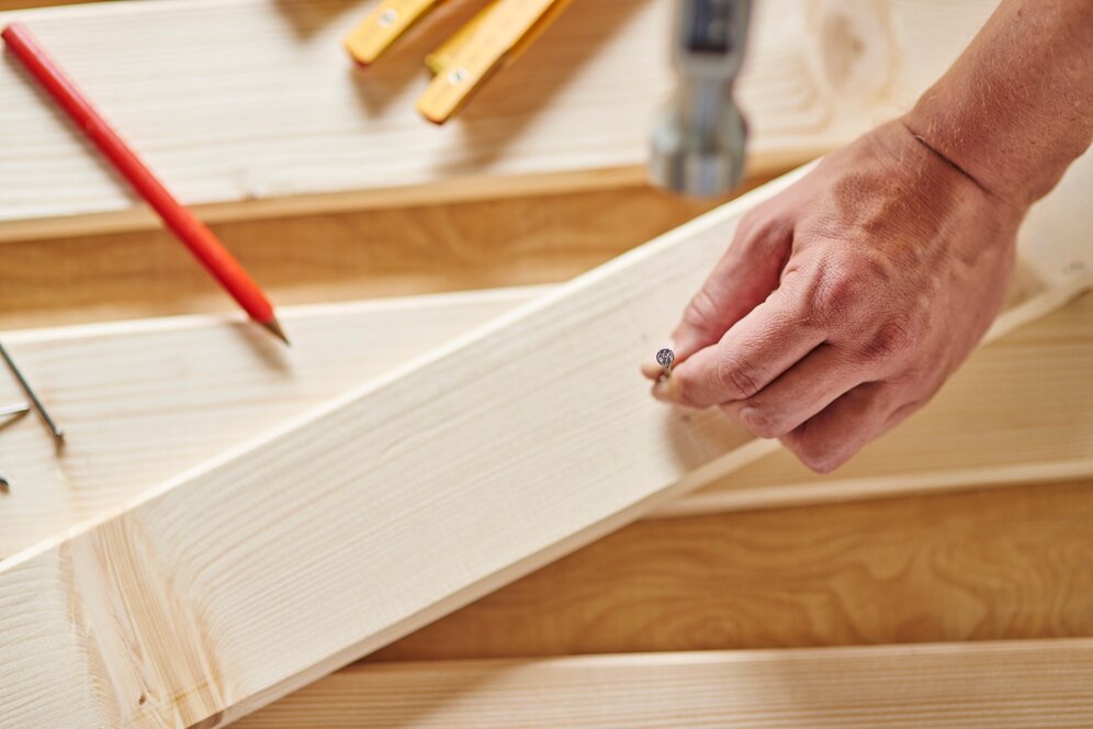 A close-up of a hand holding a nail, preparing to fasten it into a wooden plank, with tools and a red pencil nearby.
