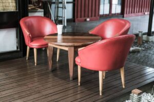 Two red leather armchairs flank a round wooden table, with a white cup on top, set on a wooden deck in a casual indoor space.
