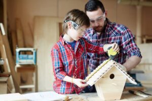 A child and adult work together to build a wooden birdhouse, using a measuring tape in a cozy workshop filled with tools.