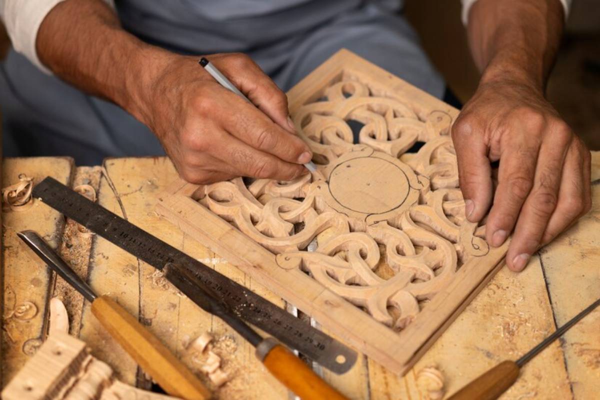 A craftsman sketches intricate designs on a carved wooden panel, surrounded by carving tools and shavings in a workshop setting.