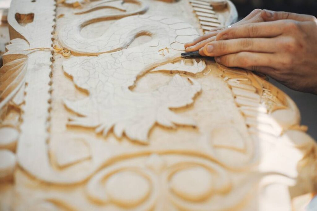 Close-up of a woodworker's hands carving intricate designs into a detailed wooden panel, showcasing craftsmanship and artistry.