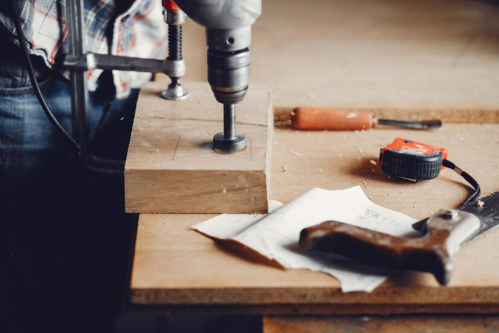 A carpenter drills into a wooden block, surrounded by tools like a measuring tape, saw, and a chisel on a workbench.