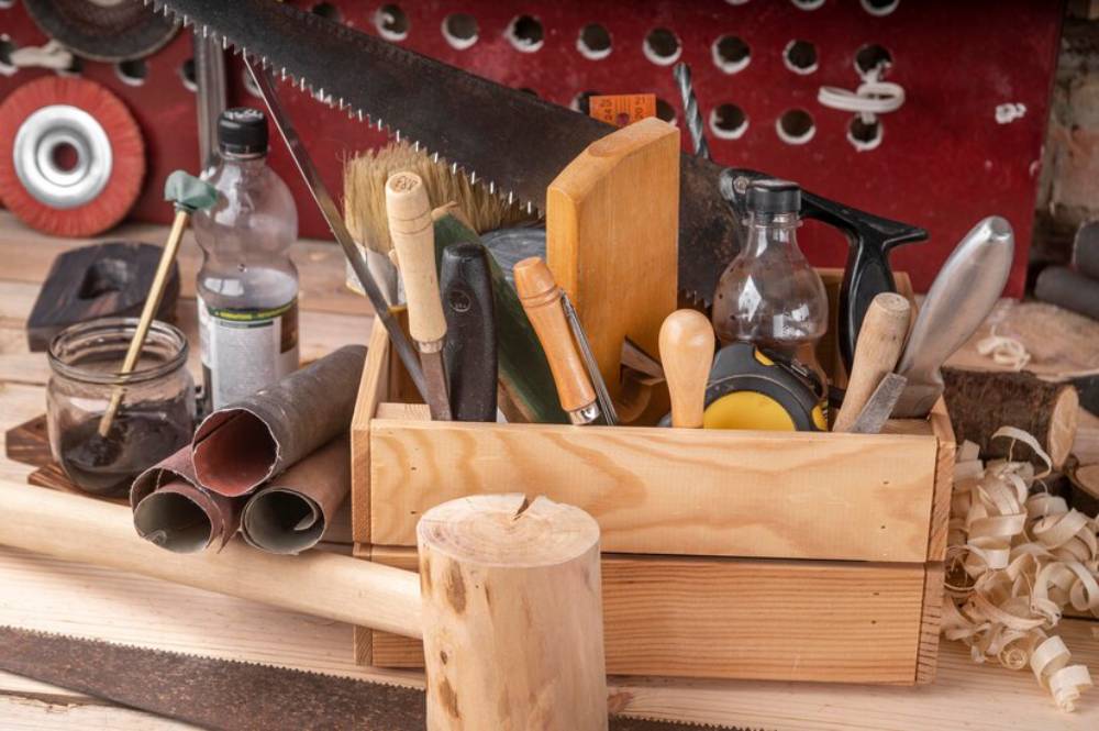 A wooden toolbox filled with various hand tools, brushes, and sandpaper, on a workshop table with wood shavings scattered around.