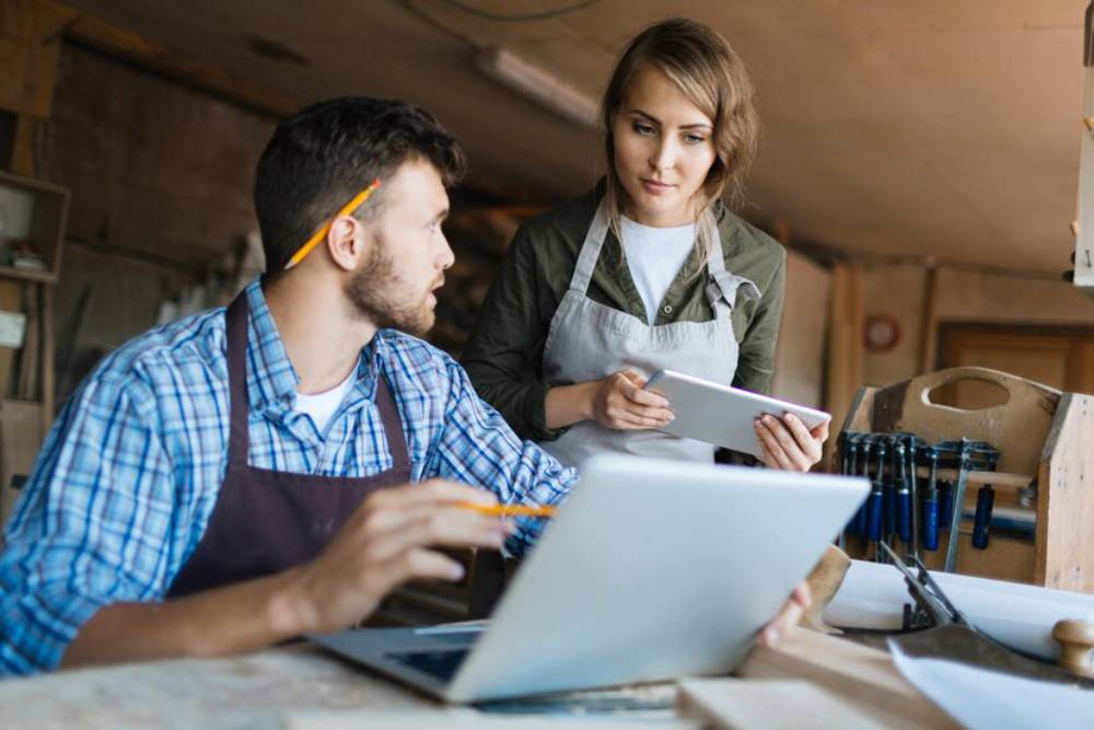 A man and a woman are collaborating in a workshop, surrounded by tools and a laptop, planning tha business using a tablet.