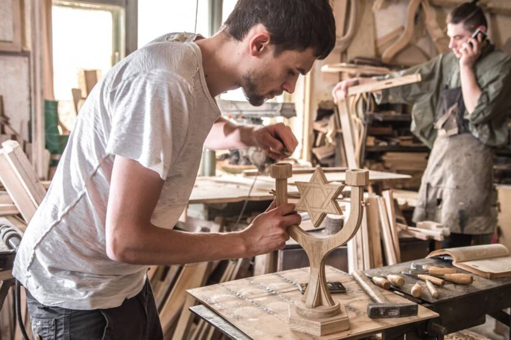 A carpenter carves a wooden menorah in a workshop filled with tools and materials.