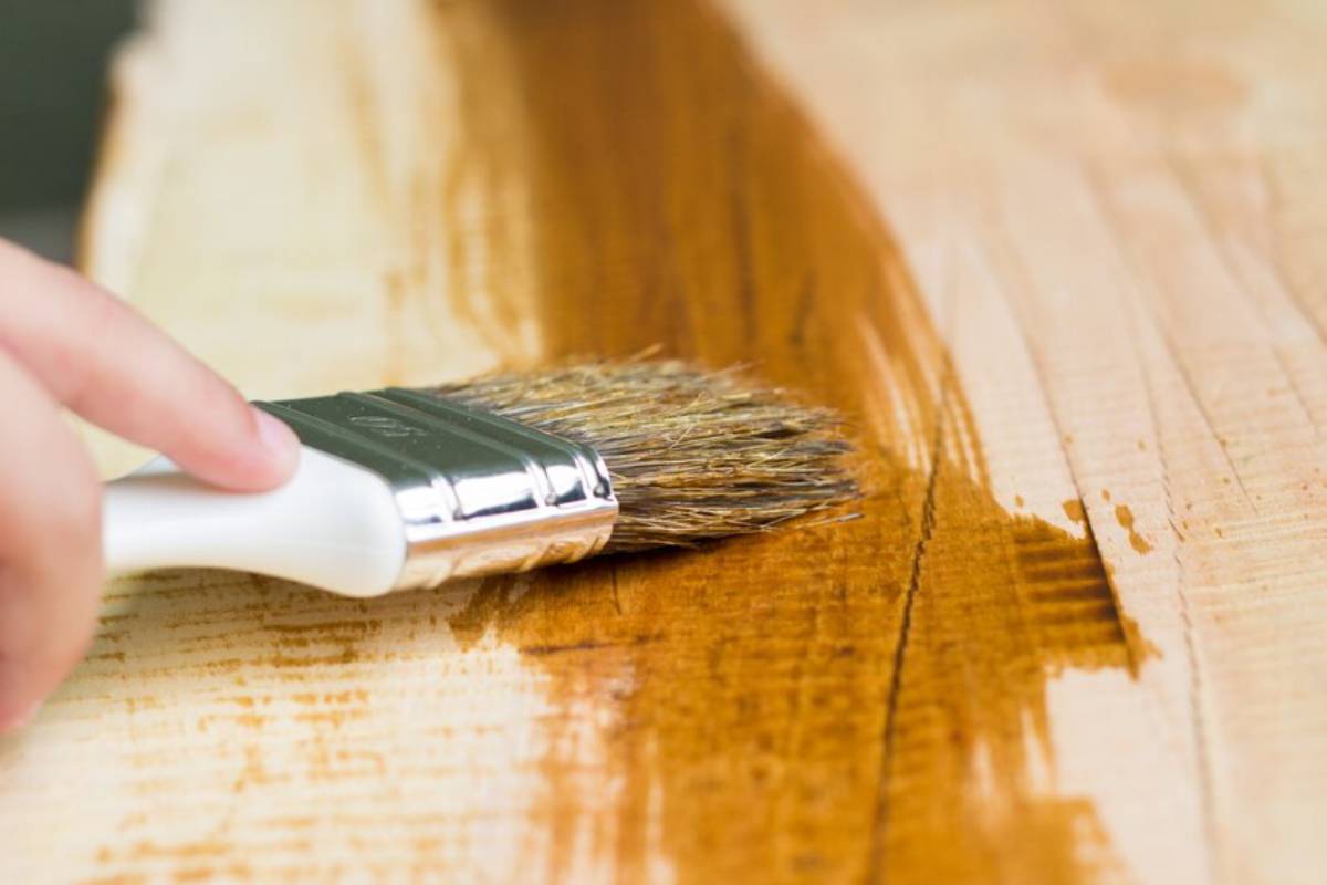 A person applies varnish with a brush to a wooden surface, giving wood finish.