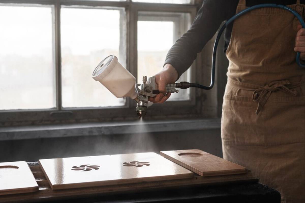 A person in an apron sprays a woodwork piece with a spray gun, surrounded by mist, near a sunlit window.