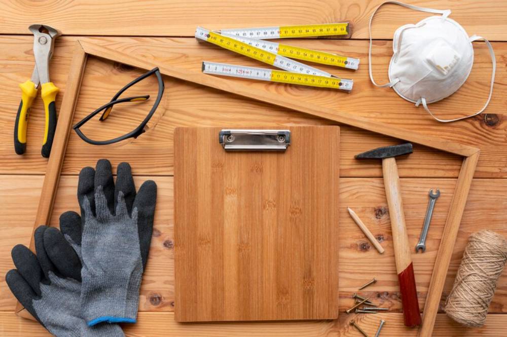 A flat lay of various DIY tools: pliers, measuring tapes, gloves, a clipboard, glasses, a hammer, a mask, and twine on a wooden surface.