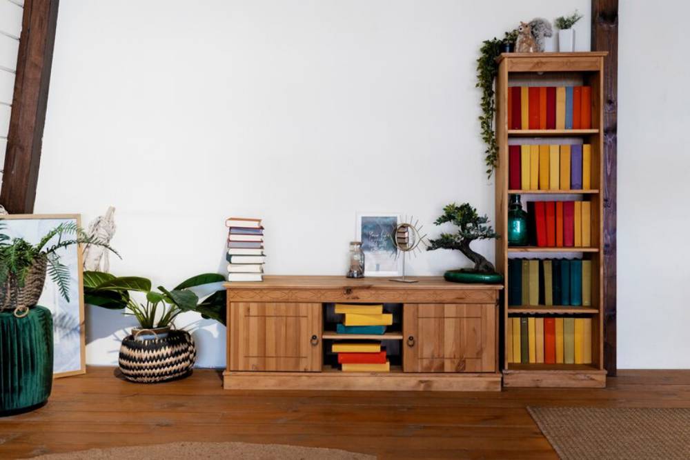 A cozy living space featuring wooden furniture, colorful bookshelves, plants, and a decorative mirror on a light-filled wall.