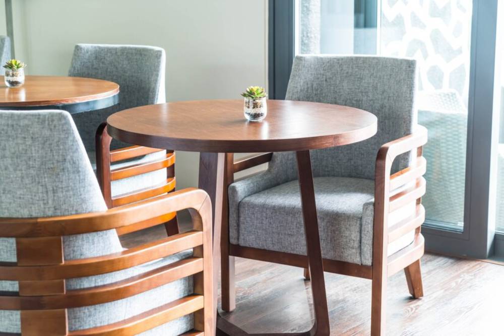 A cozy dining area featuring round wooden tables, gray upholstered chairs, and small potted succulents, by large windows.